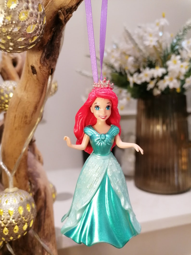May include: A red-haired princess doll wearing a turquoise gown with glitter accents. She is wearing a crown and has a purple ribbon attached to her head. The doll is hanging from a tree branch.