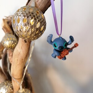 Op de afbeelding: Blauw Stitch-figuurtje ornament dat gitaar speelt, hangend aan een paars lint aan een tak met gouden kerstverlichting.