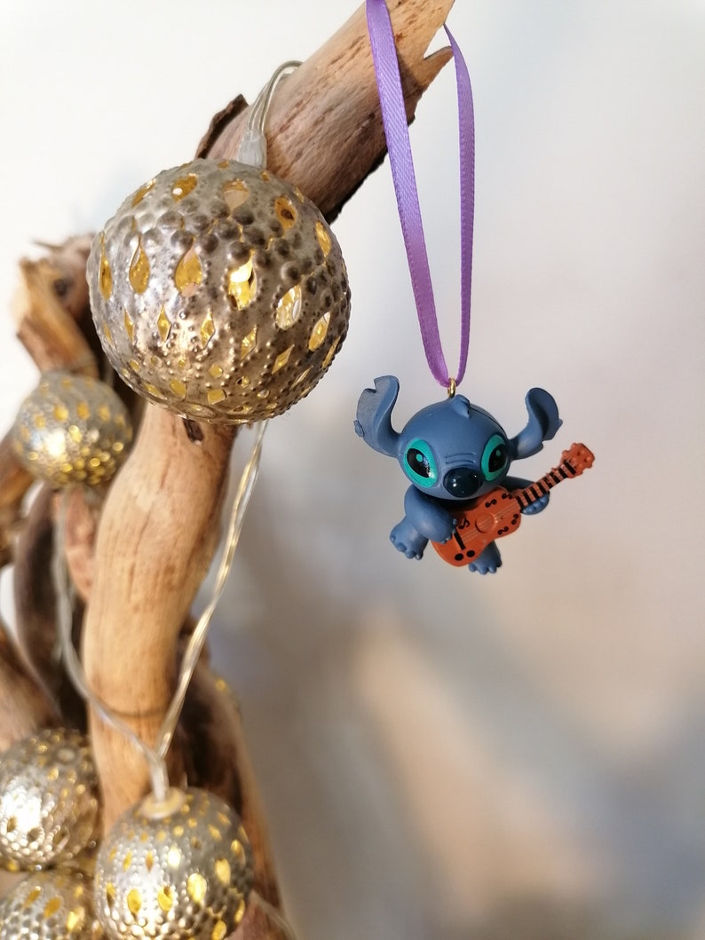 Op de afbeelding: Blauw Stitch-figuurtje ornament dat gitaar speelt, hangend aan een paars lint aan een tak met gouden kerstverlichting.