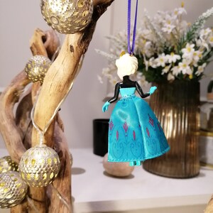 K&ouml;nnte beinhalten: Ein blauer und wei&szlig;er Plastik-Anh&auml;nger einer Prinzessin, die ein blaues Kleid mit einem wei&szlig;en Oberteil und einer Krone tr&auml;gt. Der Anh&auml;nger h&auml;ngt an einem lila Band.
