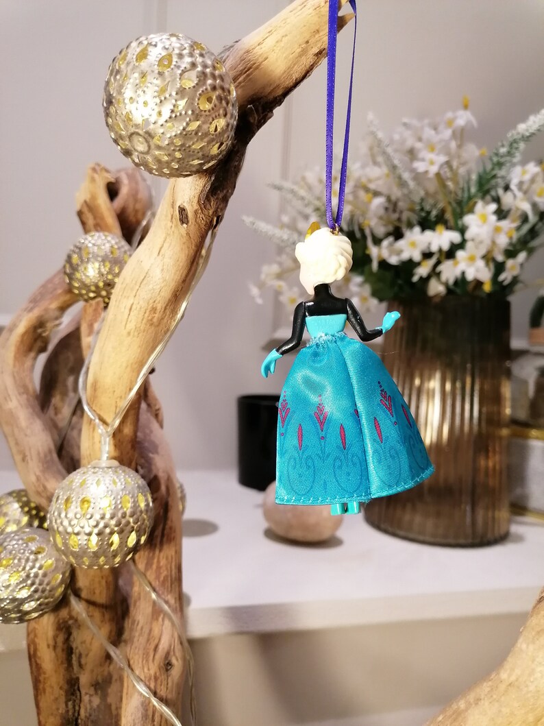 K&ouml;nnte beinhalten: Ein blauer und wei&szlig;er Plastik-Anh&auml;nger einer Prinzessin, die ein blaues Kleid mit einem wei&szlig;en Oberteil und einer Krone tr&auml;gt. Der Anh&auml;nger h&auml;ngt an einem lila Band.