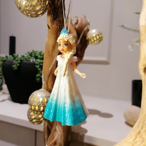 Peut inclure: Une figurine de princesse en plastique blanche et bleue, portant une robe blanche avec une jupe bleu d&eacute;grad&eacute;. La poup&eacute;e est suspendue &agrave; une branche avec une ficelle.