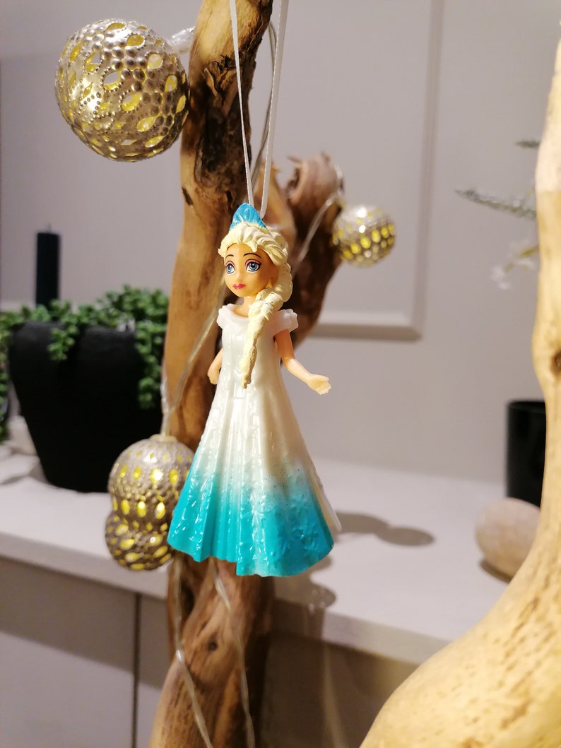 Peut inclure: Une figurine de princesse en plastique blanche et bleue, portant une robe blanche avec une jupe bleu d&eacute;grad&eacute;. La poup&eacute;e est suspendue &agrave; une branche avec une ficelle.