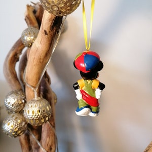 May include: A colourful Christmas bauble featuring a cartoon character wearing a red, blue and yellow hat. The character is wearing a green and yellow outfit with a red sash. The bauble is hanging from a yellow string.