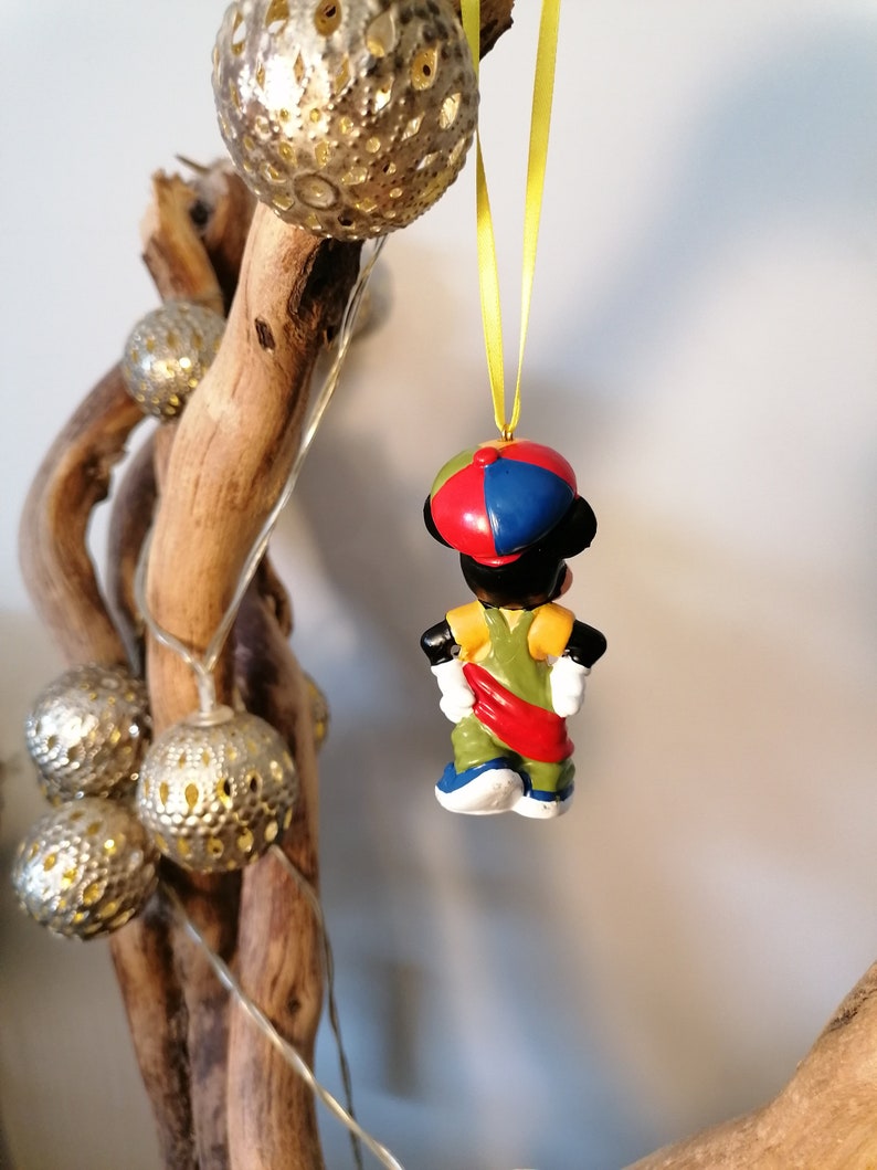 May include: A colourful Christmas bauble featuring a cartoon character wearing a red, blue and yellow hat. The character is wearing a green and yellow outfit with a red sash. The bauble is hanging from a yellow string.