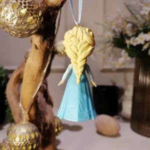 May include: A blue and white ornament of a girl with blonde hair in a braid. The ornament is hanging from a blue ribbon.