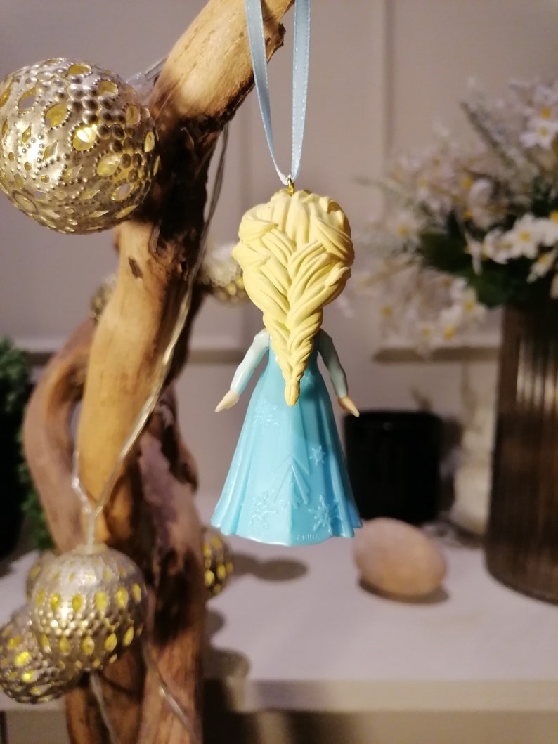 May include: A blue and white ornament of a girl with blonde hair in a braid. The ornament is hanging from a blue ribbon.
