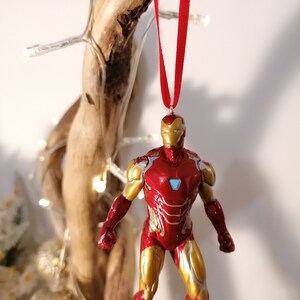 May include: Red and gold Iron Man Christmas ornament hanging from a red ribbon. The ornament is a detailed figurine of Iron Man in his red and gold suit.