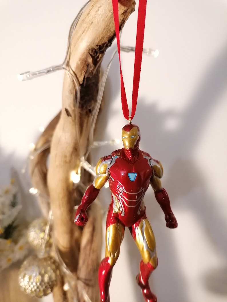 May include: Red and gold Iron Man Christmas ornament hanging from a red ribbon. The ornament is a detailed figurine of Iron Man in his red and gold suit.