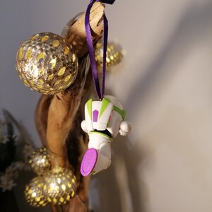 Puede incluir: Un adorno de juguete de pl&aacute;stico blanco y verde de Buzz Lightyear de la franquicia Toy Story colgado de una cinta morada en una rama con bolas doradas de brillantina.