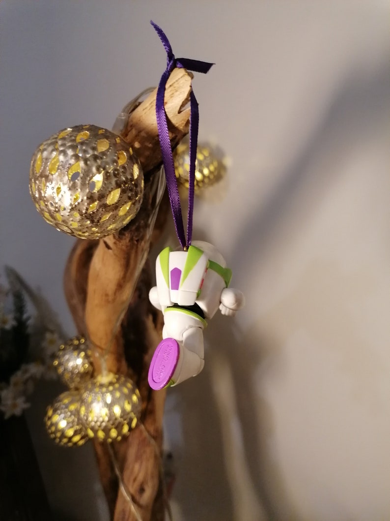 Puede incluir: Un adorno de juguete de pl&aacute;stico blanco y verde de Buzz Lightyear de la franquicia Toy Story colgado de una cinta morada en una rama con bolas doradas de brillantina.