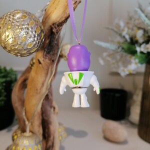 Puede incluir: Un adorno navide&ntilde;o de Buzz Lightyear blanco y verde con una cabeza morada y una cinta verde colgando de una rama.
