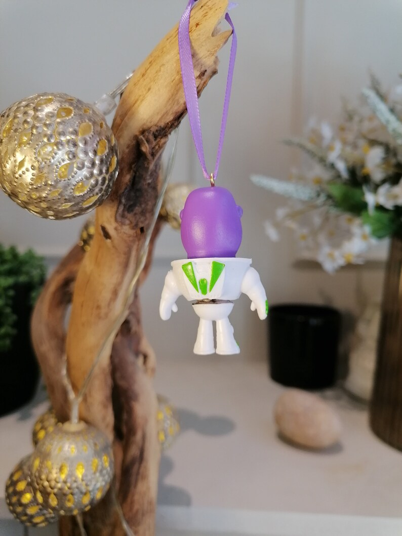 Puede incluir: Un adorno navide&ntilde;o de Buzz Lightyear blanco y verde con una cabeza morada y una cinta verde colgando de una rama.