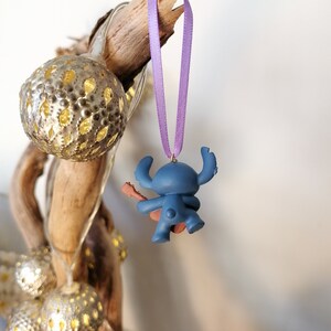 Op de afbeelding: Blauw Stitch-figuurtje ornament met een paars lint dat aan een tak hangt. Het figuurtje houdt een gitaar vast en draagt een rode gitaarband.