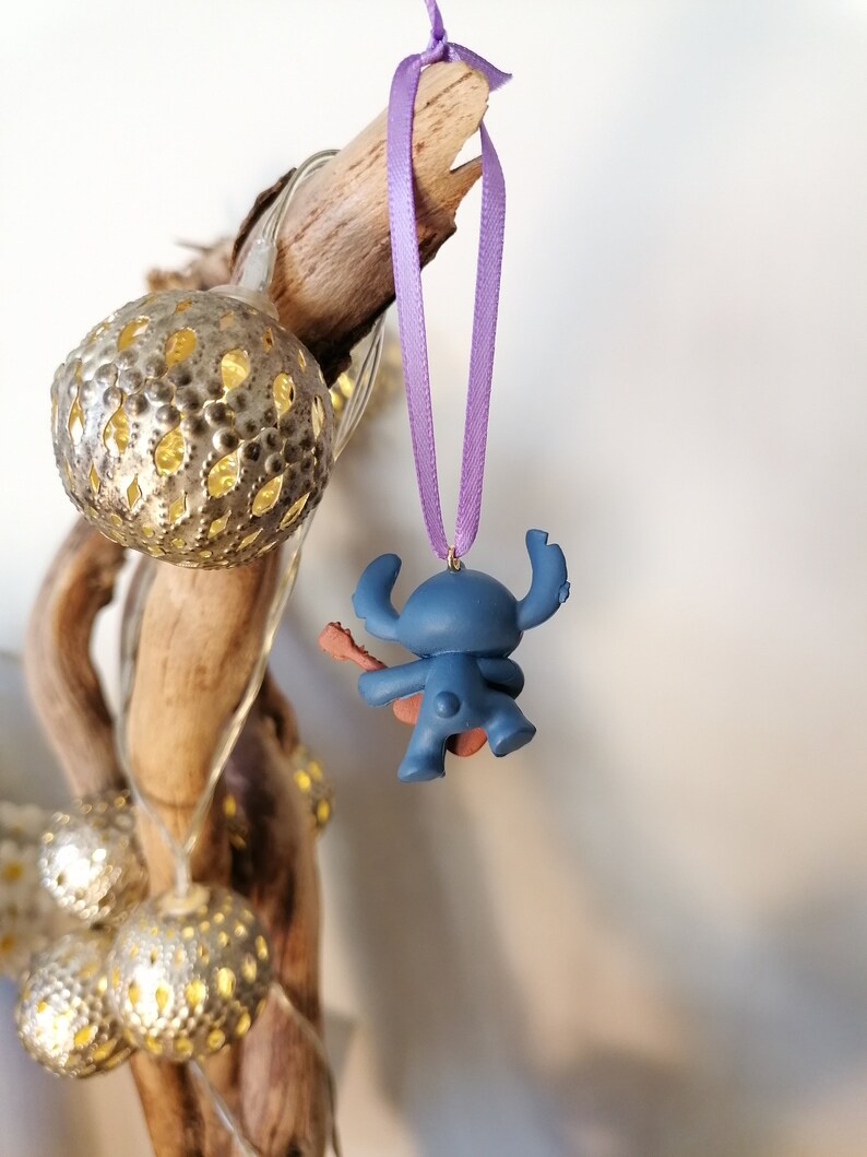 Op de afbeelding: Blauw Stitch-figuurtje ornament met een paars lint dat aan een tak hangt. Het figuurtje houdt een gitaar vast en draagt een rode gitaarband.