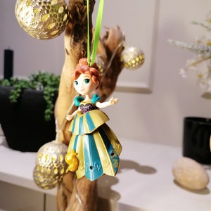 May include: A green string hangs from a branch with a gold and blue princess ornament. The ornament is a small plastic figurine of a princess with a crown and a long dress. The princess is wearing a blue and gold dress with a green sash. The ornament is hanging from a branch with a green string.