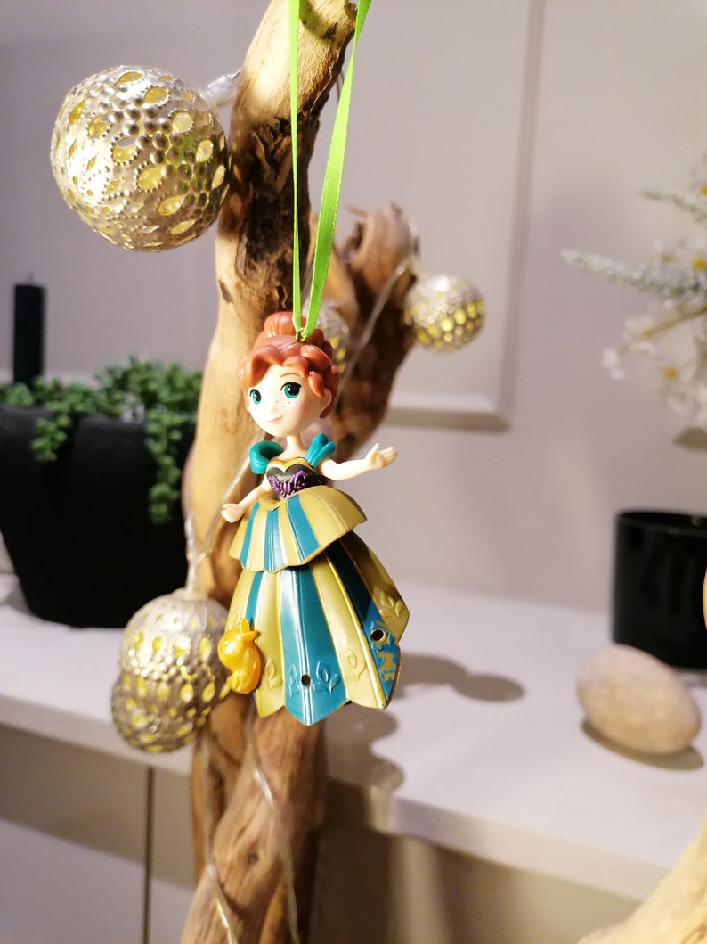 May include: A green string hangs from a branch with a gold and blue princess ornament. The ornament is a small plastic figurine of a princess with a crown and a long dress. The princess is wearing a blue and gold dress with a green sash. The ornament is hanging from a branch with a green string.