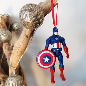 May include: A red, white, and blue Captain America action figure ornament with a red ribbon hanging from a branch. The figure is holding a red, white, and blue shield.
