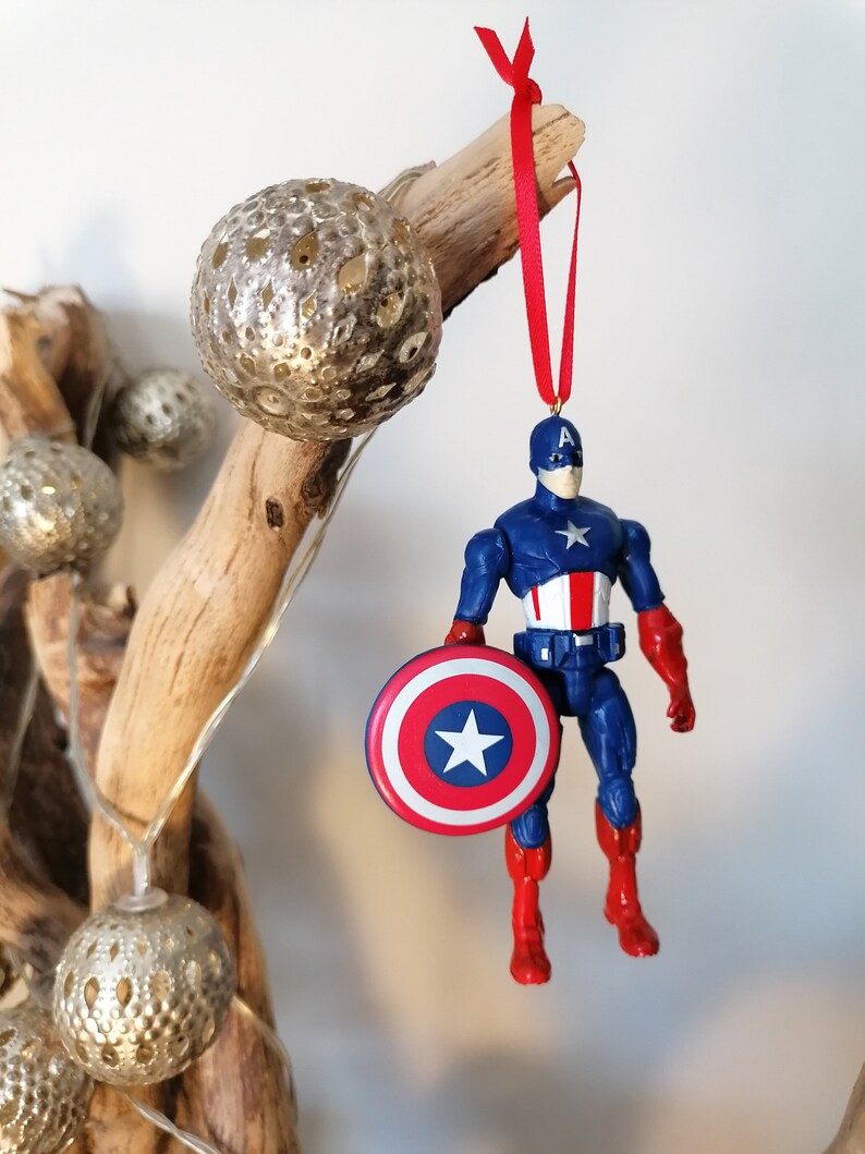 May include: A red, white, and blue Captain America action figure ornament with a red ribbon hanging from a branch. The figure is holding a red, white, and blue shield.