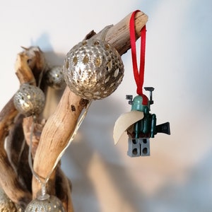 May include: A green and gray Lego minifigure of Boba Fett, wearing a tan cape, hanging from a branch with a red ribbon. The minifigure is holding a blaster rifle and has a jetpack on its back.