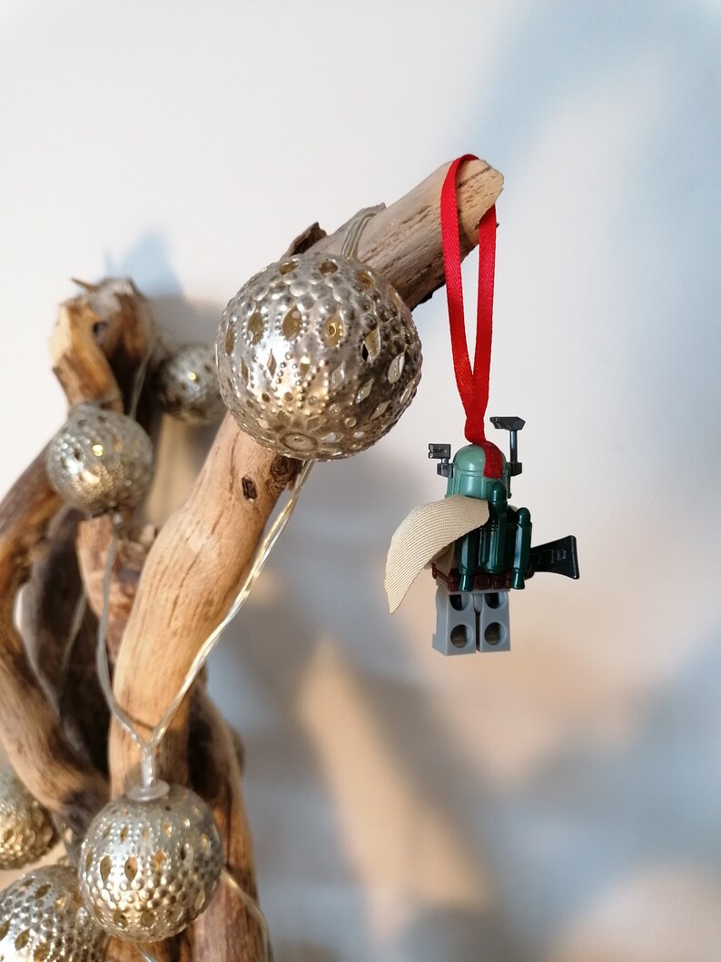 May include: A green and gray Lego minifigure of Boba Fett, wearing a tan cape, hanging from a branch with a red ribbon. The minifigure is holding a blaster rifle and has a jetpack on its back.