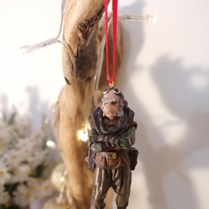 K&ouml;nnte beinhalten: Detaillierte Figur einer Figur mit Brille, Schal und milit&auml;rischem Outfit, die an einem roten Band h&auml;ngt. Die Figur hat ein verwittertes Aussehen mit Braun-, Gr&uuml;n- und Beiget&ouml;nen. Der Schmuck h&auml;ngt vor einem nat&uuml;rlichen Holzuntergrund.