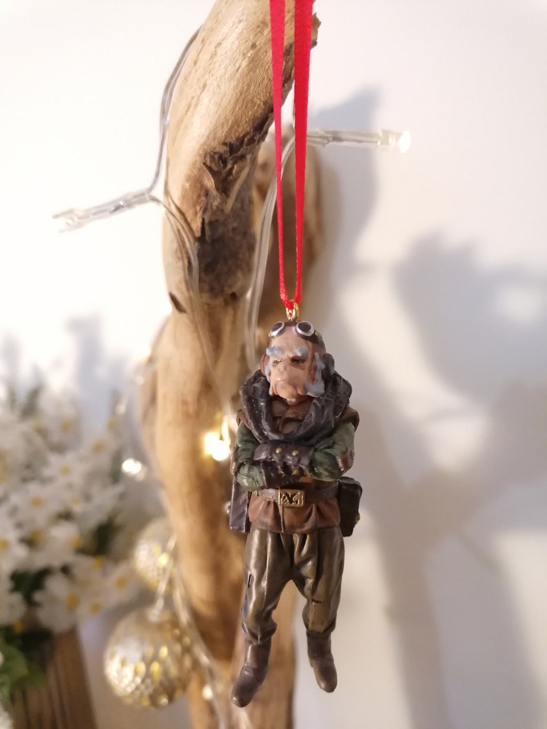 K&ouml;nnte beinhalten: Detaillierte Figur einer Figur mit Brille, Schal und milit&auml;rischem Outfit, die an einem roten Band h&auml;ngt. Die Figur hat ein verwittertes Aussehen mit Braun-, Gr&uuml;n- und Beiget&ouml;nen. Der Schmuck h&auml;ngt vor einem nat&uuml;rlichen Holzuntergrund.