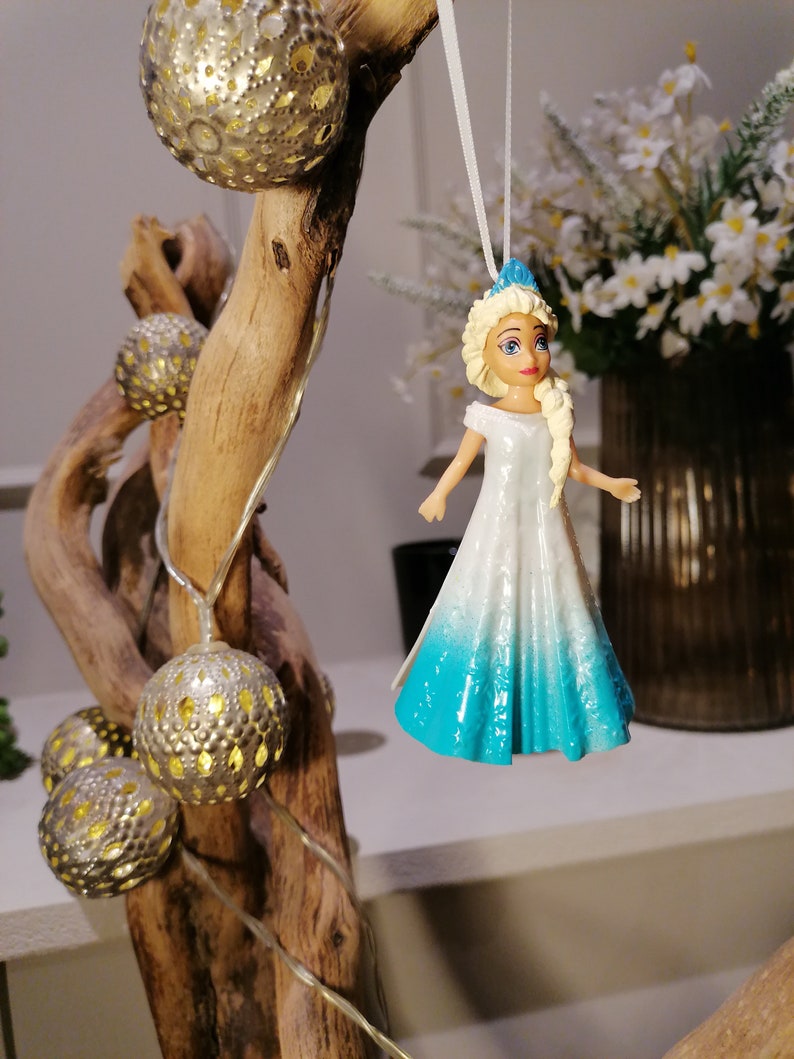 Peut inclure: Une figurine de princesse blanche et bleue avec des cheveux blonds et une couronne bleue. La poup&eacute;e est suspendue &agrave; une ficelle blanche sur une branche avec des ampoules dor&eacute;es.