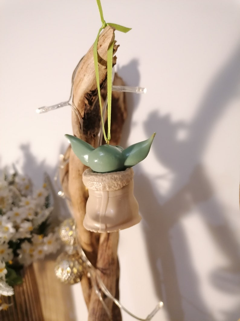 May include: A green and brown Christmas ornament featuring a character from the Star Wars franchise. The ornament is hanging from a green ribbon.