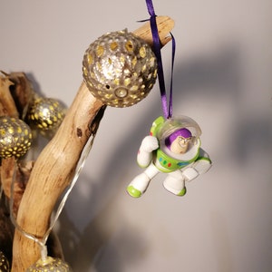 Puede incluir: Adorno navideño con Buzz Lightyear en su traje espacial blanco, verde y morado, colgado de una cinta morada. También se observa un adorno esférico plateado con diseños recortados.