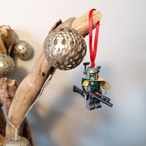 May include: A green and brown Lego Star Wars Boba Fett minifigure ornament hanging from a branch with a red ribbon. The ornament is attached to a silver and white decorative ball.