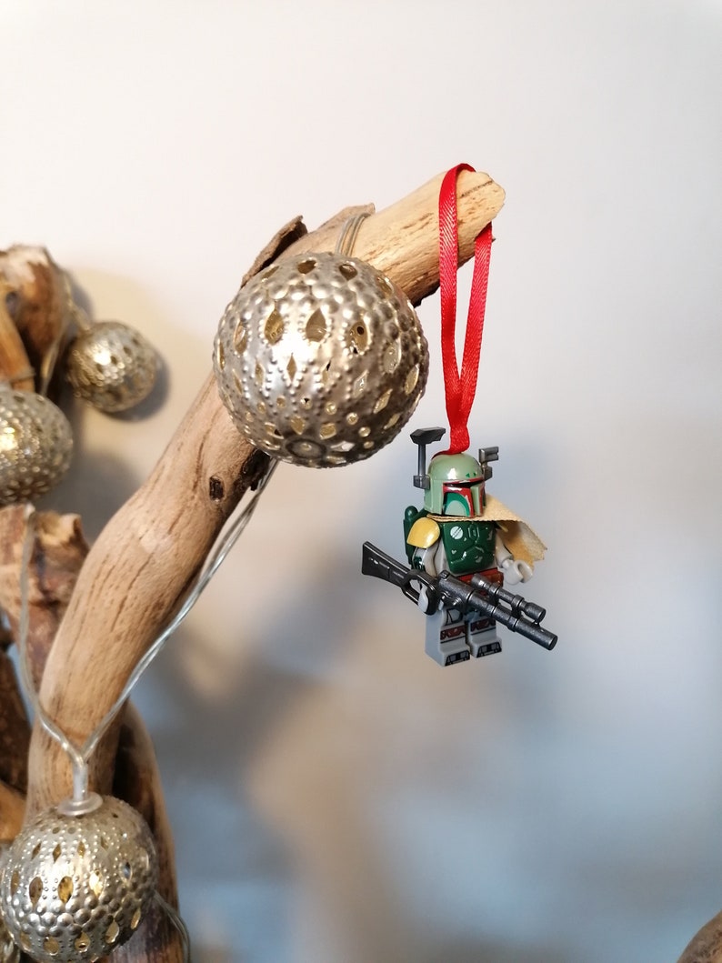 May include: A green and brown Lego Star Wars Boba Fett minifigure ornament hanging from a branch with a red ribbon. The ornament is attached to a silver and white decorative ball.