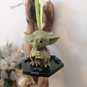 Puede incluir: Adorno de Navidad de Star Wars Yoda verde con una base hexagonal negra y una cinta verde.