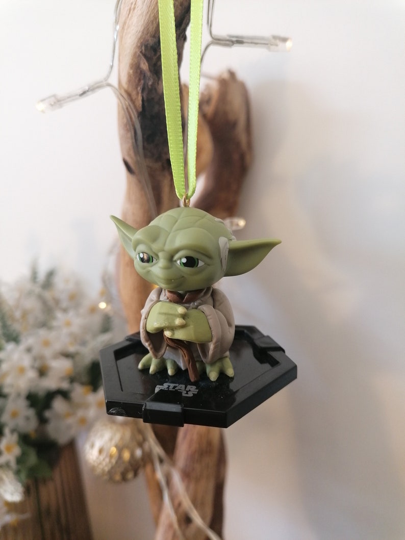 Puede incluir: Adorno de Navidad de Star Wars Yoda verde con una base hexagonal negra y una cinta verde.