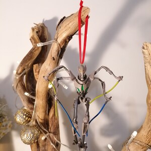 May include: A gray and green robot ornament hanging from a branch with a red ribbon. The robot has multiple arms and legs and is holding two lightsabers.