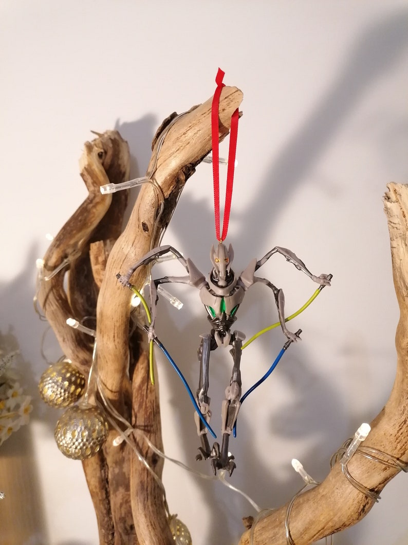 May include: A gray and green robot ornament hanging from a branch with a red ribbon. The robot has multiple arms and legs and is holding two lightsabers.