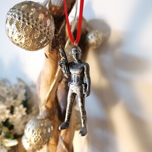 May include: A silver-coloured ornament of a female figure holding a weapon. The ornament is hanging from a red ribbon.