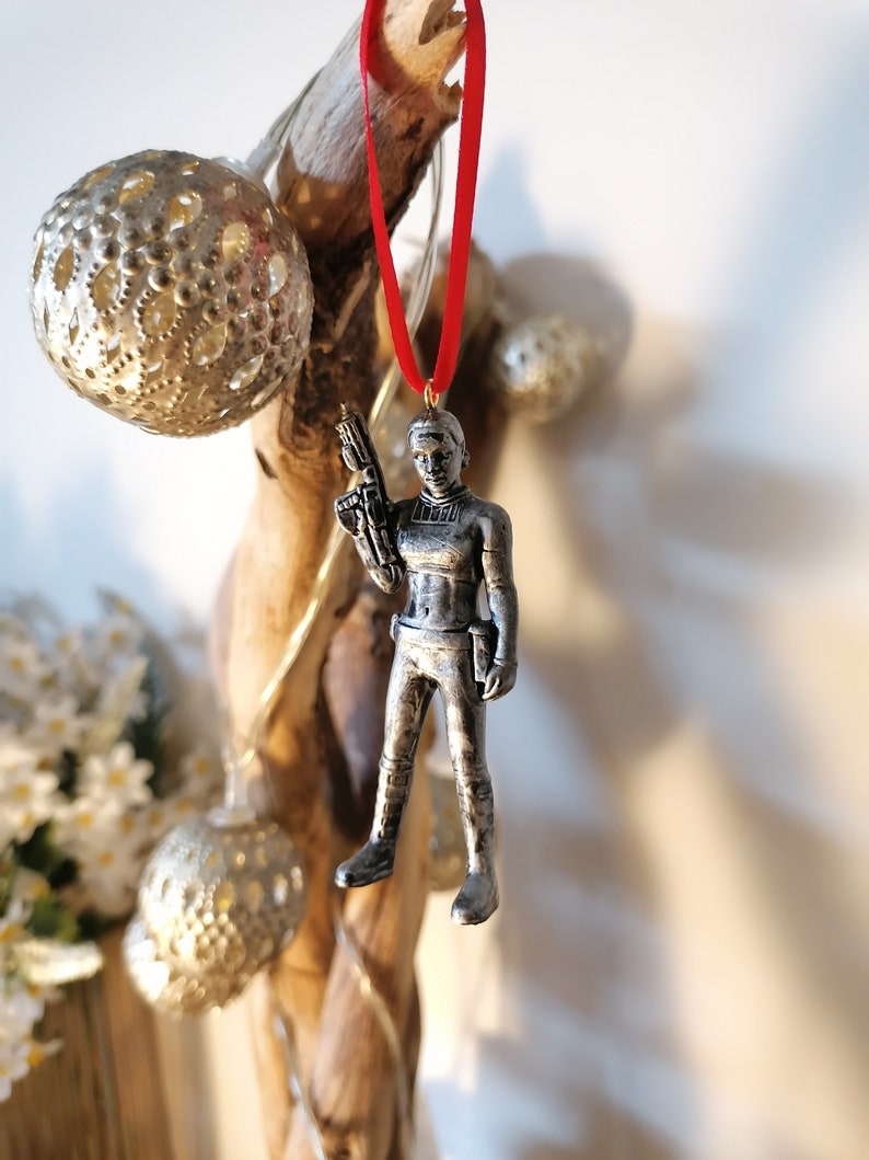 May include: A silver-coloured ornament of a female figure holding a weapon. The ornament is hanging from a red ribbon.
