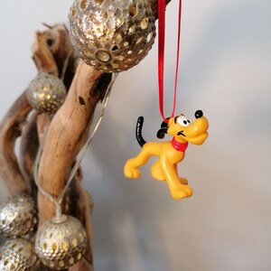 Puede incluir: Una figurita de plástico amarilla y negra de Pluto el perro colgando de una cinta roja. La figurita está unida a una rama de madera flotante con adornos plateados y dorados.