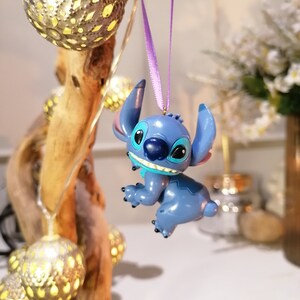 Pode incluir: Ornamento de personagem Stitch azul pendurado em uma fita roxa. O ornamento &eacute; uma figura 3D do personagem com um corpo azul, olhos brancos e uma boca azul. O ornamento est&aacute; pendurado em um galho com luzes brancas.