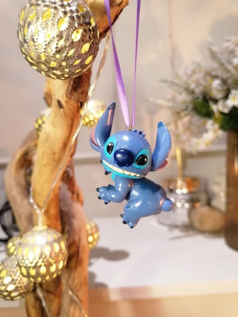 Pode incluir: Ornamento de personagem Stitch azul pendurado em uma fita roxa. O ornamento &eacute; uma figura 3D do personagem com um corpo azul, olhos brancos e uma boca azul. O ornamento est&aacute; pendurado em um galho com luzes brancas.