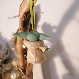 May include: A green and brown Christmas ornament shaped like a character from the Star Wars franchise. The ornament is hanging from a green ribbon.