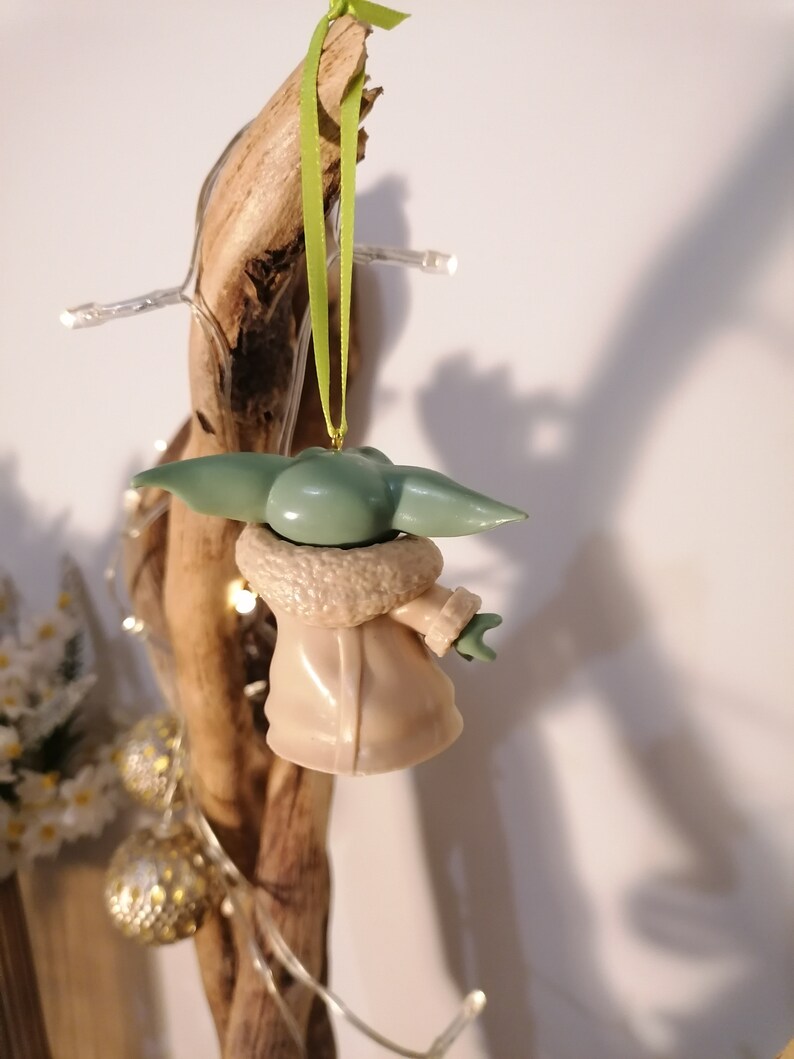May include: A green and brown Christmas ornament shaped like a character from the Star Wars franchise. The ornament is hanging from a green ribbon.