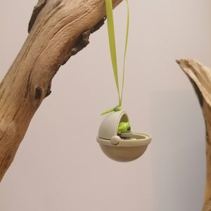 May include: A small, tan-colored toy pod with a green figure inside, suspended by a light green ribbon. The pod is hanging from a piece of driftwood, set against a neutral background. The toy is a collectible item.