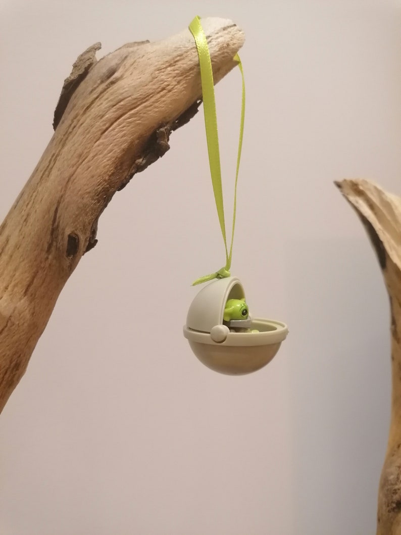 May include: A small, tan-colored toy pod with a green figure inside, suspended by a light green ribbon. The pod is hanging from a piece of driftwood, set against a neutral background. The toy is a collectible item.