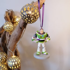 May include: A green and white Buzz Lightyear Christmas ornament with a purple ribbon hanging from a branch with string lights.