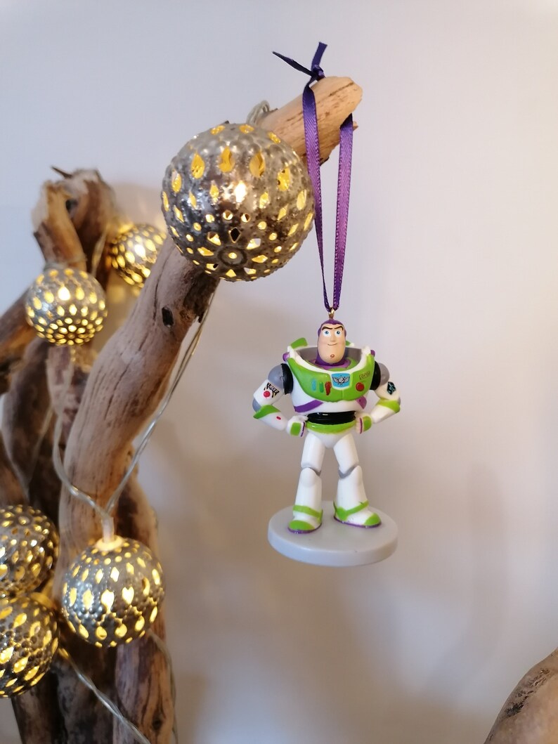 May include: A green and white Buzz Lightyear Christmas ornament with a purple ribbon hanging from a branch with string lights.