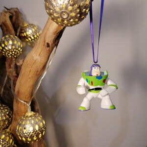 May include: A green and white plastic toy ornament of Buzz Lightyear from the Toy Story movie franchise. The ornament is hanging from a purple ribbon on a branch with gold decorative lights.