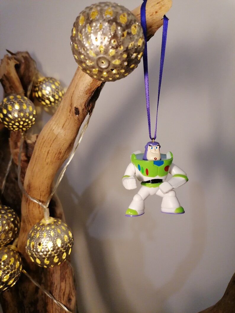 May include: A green and white plastic toy ornament of Buzz Lightyear from the Toy Story movie franchise. The ornament is hanging from a purple ribbon on a branch with gold decorative lights.