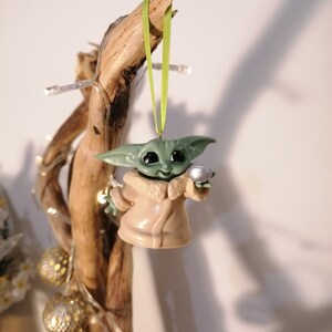 May include: A green and brown Christmas ornament featuring a character from the Star Wars franchise, holding a silver bell. The ornament is hanging from a green ribbon on a branch.